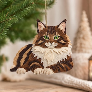 Può includere: Un ornamento a forma di gatto in feltro con un design Maine Coon tigrato marrone e crema, occhi verdi, dettagli cuciti e un cordino dorato. Il gatto è raffigurato in posizione di riposo con un folto pelo sul petto e orecchie a ciuffi.