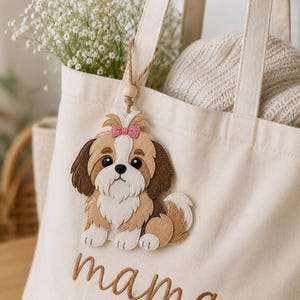 May include: Cream-colored tote bag with the word "mama" embroidered in brown script, featuring a felt Shih Tzu dog applique with a pink bow. The bag has a wooden bead charm and is filled with flowers and a sweater.
