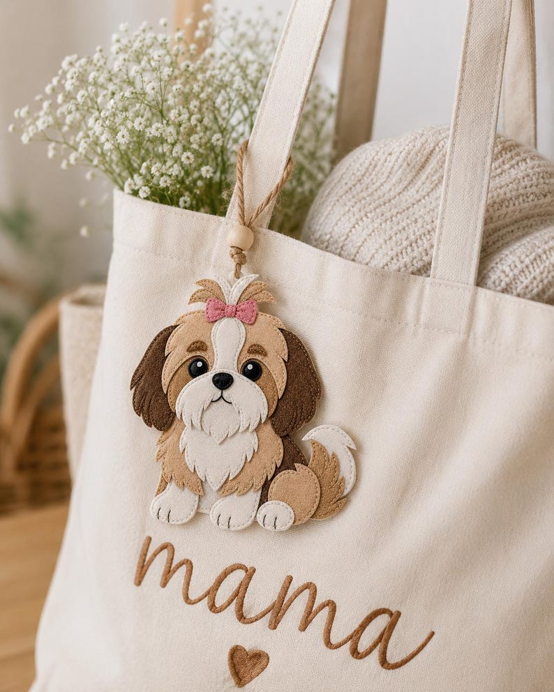 May include: Cream-colored tote bag with the word "mama" embroidered in brown script, featuring a felt Shih Tzu dog applique with a pink bow. The bag has a wooden bead charm and is filled with flowers and a sweater.