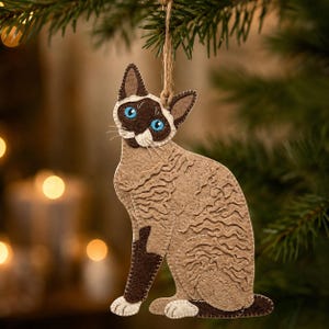 Può includere: Un ornamento in feltro a forma di gatto siamese con corpo color marrone chiaro e punte marrone scuro su orecchie, viso e zampe. Il gatto ha occhi azzurri brillanti, zampe bianche e una superficie testurizzata. È appeso a un sottile cordino di juta.