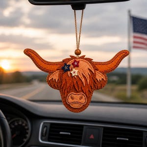 Op de afbeelding: Een vilten Highland koe auto-ornament in bruin- en oranjetinten. De charme toont een koeienkop met hoorns, een ruige manen en sterdecoraties in rood, wit en blauw. Het hangt aan een touwtje met een houten kraal.