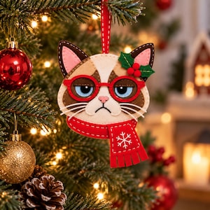 Puede incluir: Un adorno de fieltro con forma de gato de expresión gruñona, que lleva gafas rojas y una bufanda roja a juego con un diseño de copo de nieve blanco. El gato tiene pelaje marrón y crema, orejas rosas y un adorno de acebo verde con bayas.