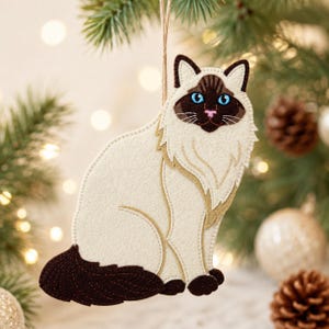 May include: A felt cat ornament featuring a cream-colored body with dark brown ears, face, and tail. The cat has bright blue eyes, a pink nose, and detailed stitching throughout. It hangs from a thin twine string, showcasing a layered design.