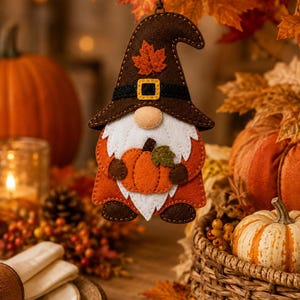 May include: A felt gnome ornament featuring a brown pointed hat with a stitched orange maple leaf and a black band with a yellow buckle. The gnome has a white beard, an orange coat, brown boots, and holds a small orange pumpkin with a green leaf.