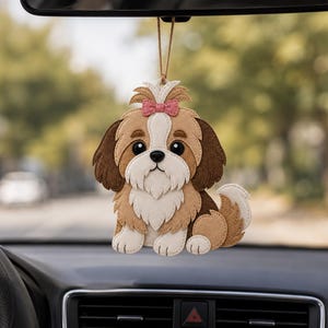 May include: A felt Shih Tzu car charm with a pink bow. The dog has brown, tan, and white fur, with large black eyes. The charm hangs from a string, and is displayed in a car interior.