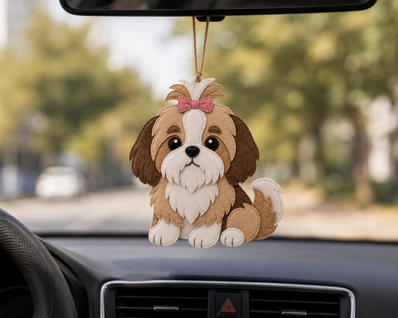 May include: A felt Shih Tzu car charm with a pink bow. The dog has brown, tan, and white fur, with large black eyes. The charm hangs from a string, and is displayed in a car interior.