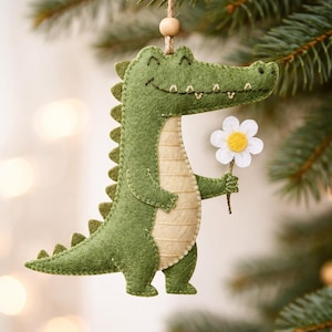 Può includere: Ornamento natalizio fatto a mano in feltro verde a forma di alligatore che tiene una margherita bianca con centro giallo. L'alligatore ha la pancia color crema e un viso sorridente. L'ornamento è sospeso da uno spago.