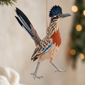 May include: A felt roadrunner ornament featuring a tan, brown, and rust-colored body with a blue-spotted crest, a long black and grey tail, and grey legs. The bird is suspended by a thin twine string, showcasing detailed stitching and layered fabric.