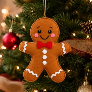 May include: A brown gingerbread person ornament featuring a red bow tie, pink cheeks, and white icing details on the arms and legs. The figure has black eyes and a stitched smile, suspended by a gold string for hanging on a holiday tree.