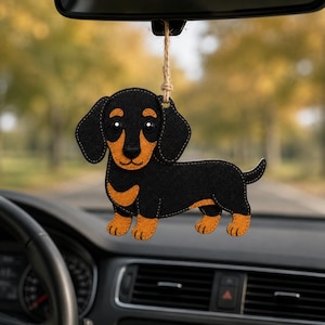 May include: A felt dachshund car charm. The black and tan dog is hanging from a rearview mirror. The dog has a black body, tan paws, and a tan face. The charm is hanging in front of a car dashboard.