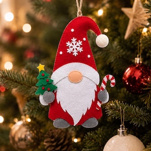 May include: A felt Christmas gnome ornament featuring a red hat with a white snowflake and pom-pom, a white beard, a peach-colored nose, and grey feet. The gnome holds a green Christmas tree with a yellow star and a red and white candy cane.