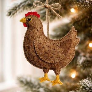 Può includere: Un ornamento a forma di gallina in feltro marrone con cresta rossa e becco e zampe gialle. L'ornamento è appeso a uno spago legato a fiocco. Lo sfondo è un'immagine sfocata di un albero di Natale.