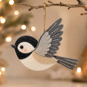 Op de afbeelding: Een vilten vogelornament met een zwarte kop, grijze vleugels en een beige lichaam, hangend aan een tak. De vogel heeft een kleine, roze wang en een wit oog. De vleugels zijn gedetailleerd met witte stiksels. De achtergrond bevat een vage kerstboom en lichten.
