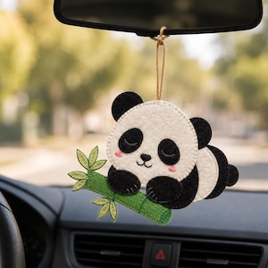 May include: A felt hanging ornament shaped like a sleeping panda bear holding a green bamboo stalk. The panda is white with black ears, eye patches, and limbs, featuring pink cheeks and a brown string loop for hanging from a rearview mirror.