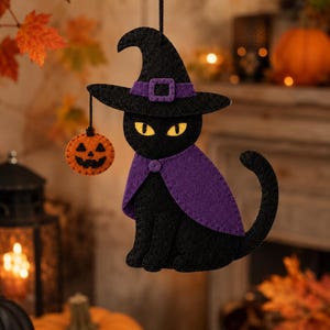Puede incluir: Un adorno de fieltro negro con forma de gato que lleva una capa morada y un sombrero de bruja a juego con una hebilla morada. El gato tiene ojos amarillos y sostiene una pequeña calabaza naranja en una cuerda. Cuelga de un cordón negro.