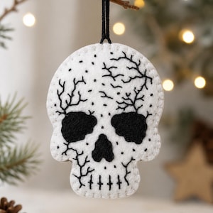 Op de afbeelding: Een witte vilt schedelornament met zwarte geborduurde details, waaronder grote oogkassen, een driehoekige neus en gestikte tanden. Het oppervlak is versierd met zwarte takachtige patronen en kleine stippen, afgewerkt met een zwart ophangkoord.