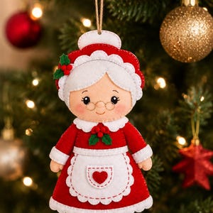 May include: A felt Christmas ornament shaped like a grandmother figure wearing a red dress with a white apron, a white bonnet, and gold-rimmed glasses. The ornament features green holly leaves with red berries and a small red heart on the apron.