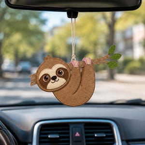 May include: A brown felt sloth hanging ornament suspended by a string. The sloth has a cream-colored face, large dark eyes, and is clutching a brown branch with three small green leaves. The design features visible stitching along the edges.