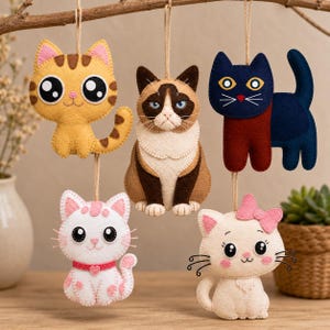 Op de afbeelding: Vijf handgemaakte vilten kattenornamenten in verschillende kleuren en ontwerpen, hangend aan een tak. De katten omvatten een gele tabby, een chagrijnige kat, een blauwe kat met een rode sjaal en twee witte katten met roze accenten.