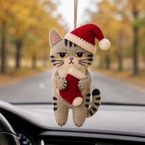 Puede incluir: Un adorno colgante de fieltro con forma de gato atigrado gris con rayas oscuras, que lleva un gorro de Papá Noel rojo y blanco y sostiene un pequeño calcetín navideño rojo y blanco. El gato tiene ojos amarillos, nariz rosa y cuelga de un hilo.