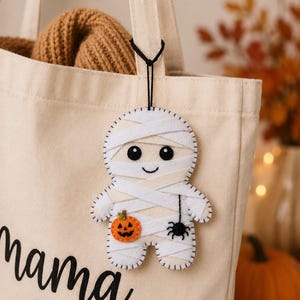 May include: A cream-coloured canvas tote bag with the word mama and a heart printed in black, featuring a hanging felt mummy ornament decorated with a small orange jack-o'-lantern and a black spider, attached by a thin black cord.