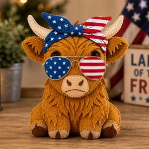 Puede incluir: Figura de fieltro de una vaca de las Tierras Altas con accesorios patrióticos. Lleva una diadema azul con estrellas blancas y gafas de sol con un diseño de rayas rojas y blancas. Un letrero de madera detrás dice "Land of the Free".