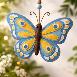 May include: A handcrafted butterfly ornament made of felt. The butterfly features blue, yellow, and white layered wings with a brown body and antennae. It hangs from a twine string with wooden beads. Decorative item.