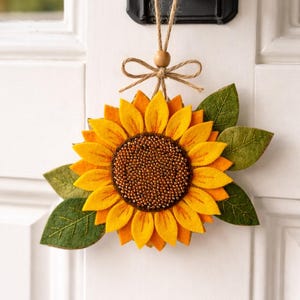 May include: A felt sunflower door decoration with a brown beaded center, yellow petals, and green leaves. The sunflower is suspended by a twine loop and wooden bead, hanging from a black door handle. A charming, handcrafted accent.
