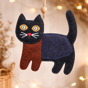May include: A handmade felt cat ornament with a black head, yellow eyes, and a red nose. The body is brown and navy blue, with a curved tail. It hangs from a natural twine string with a wooden bead.