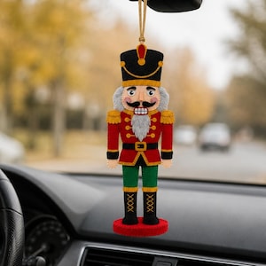 May include: A felt nutcracker ornament hanging from a rearview mirror. The figure wears a black hat, red jacket with gold trim, green trousers, and black boots. It features a white beard, mustache, and a circular red base, suspended by a gold cord.