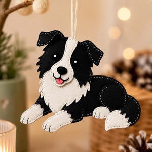 May include: A black and white felt dog ornament featuring a border collie design with a white chest, face blaze, and paws. The dog is in a lying position with its tongue visible, stitched with white thread, and suspended by a thin white string.
