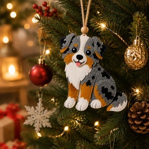 May include: A felt Australian Shepherd dog ornament featuring a gray, black, and white mottled coat with tan accents, a white chest, and a pink tongue. The ornament is suspended by a jute twine loop with a small wooden bead at the top.