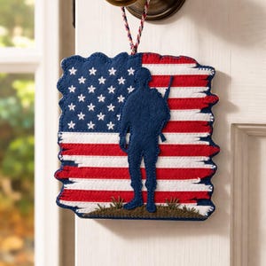 May include: A patriotic door hanger featuring the American flag with a silhouette of a soldier. The flag design includes red and white stripes, blue field with white stars, and a blue soldier silhouette. The door hanger is made of felt.