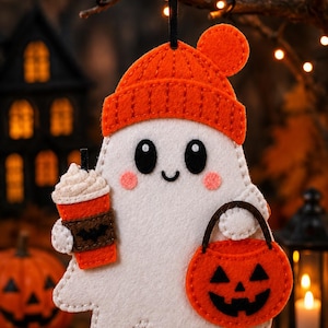 May include: A white felt ghost ornament wearing an orange beanie with a pom-pom. The ghost has black eyes, a small smile, and pink cheeks. It holds an orange coffee cup with a black bat icon and a small orange jack-o'-lantern trick-or-treat bucket.