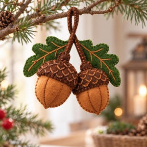 Può includere: Un ornamento in feltro raffigurante due ghiande color marrone chiaro con cappucci testurizzati marrone scuro e foglie di feltro verde con venature ricamate. Le ghiande sono collegate da un occhiello intrecciato marrone per appenderle.