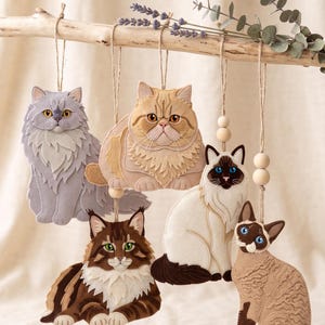 May include: Five felt cat ornaments hanging from a wooden branch. The cats include a grey long-haired cat, a tan Persian, a brown and white tabby, a white and brown Siamese-style cat, and a tan cat with dark brown points, all with twine.