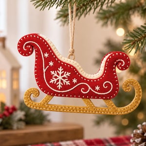 Pode incluir: Um enfeite de Natal de feltro em forma de trenó, com um corpo vermelho profundo com padrões bordados em branco de flocos de neve e espirais, uma borda cor de creme e patins dourados. O enfeite está pendurado por um cordão bege simples.
