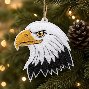 Pode incluir: Um enfeite de Natal de feltro em forma de cabeça de águia. O enfeite apresenta uma cabeça branca, um bico e um olho amarelos e penas pretas. Uma corda marrom é anexada para pendurar. O enfeite é exibido em uma árvore de Natal.
