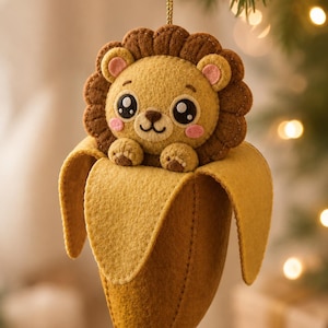 Puede incluir: Adorno navideño de fieltro con un lindo cachorro de león dentro de una cáscara de plátano. El león tiene un cuerpo marrón, una melena marrón y detalles rosas. La cáscara de plátano es amarilla. El adorno cuelga de un hilo dorado.