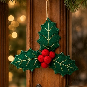 May include: A hanging ornament shaped like three dark green felt holly leaves with light green embroidered veins and a cluster of four bright red felt berries in the centre, suspended by a thin tan twine string against a wooden door frame.