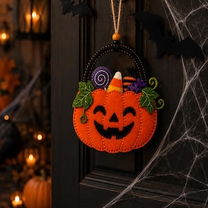 Könnte beinhalten: Eine Halloween-Dekoration aus Filz in Form eines lächelnden orangefarbenen Kürbiskorbs, gefüllt mit einem violetten Spiral-Lutscher, einem Zuckermais und einem gestreiften Bonbon. Das Ornament hat grüne Filzblätter und hängt an einem schwarzen Henkel.