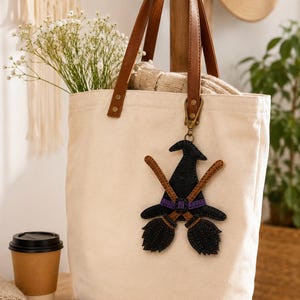 May include: A cream-colored canvas tote bag with brown leather handles, featuring a hanging bag charm shaped like a black witch hat with a purple band and two crossed brown broomsticks, attached to the bag with a gold-toned metal lobster clasp.