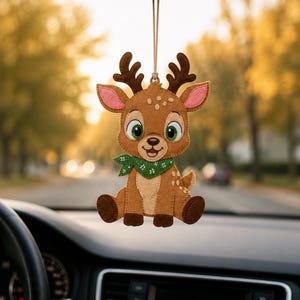 May include: A felt hanging ornament shaped like a cartoon deer with brown fur, white spots, and large green eyes. The deer wears a green bandana with a white floral pattern around its neck and has dark brown antlers, ears with pink centers, and paws.