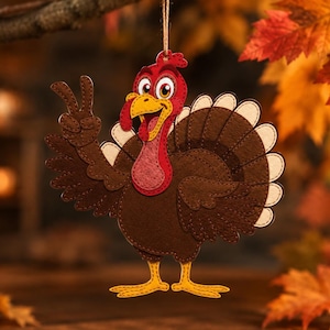 May include: A felt turkey ornament featuring a brown body, red wattle, and yellow feet. The turkey is making a peace sign with one wing and has a large, fanned-out tail with cream-colored edges. It hangs from a thin twine string at the top.