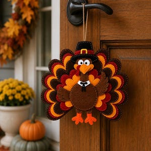 Puede incluir: Un adorno colgante de fieltro con forma de pavo que presenta un cuerpo marrón, patas naranjas y una cola de varias capas en tonos marrones, rojos y naranjas. El pavo lleva un sombrero negro con una hebilla amarilla y un cuello blanco con pajarita negra.