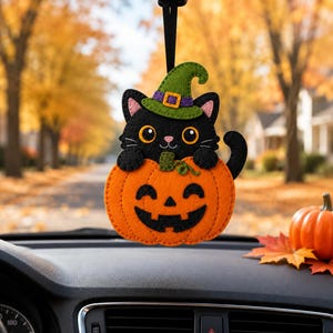 May include: A felt car air freshener hanging from a rearview mirror, featuring a black cat wearing a green witch hat with a purple band, sitting inside a bright orange jack-o'-lantern pumpkin with a black stitched face and green stem.
