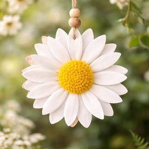 Puede incluir: Adorno de margarita blanca grande con centro amarillo brillante, suspendido por un cordón de hilo natural y cuentas de madera. La margarita tiene múltiples pétalos blancos y un fondo verde borroso.
