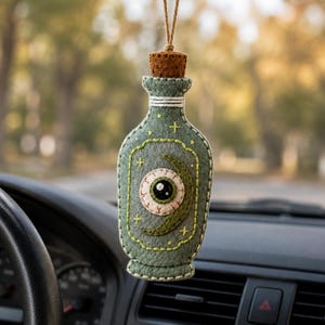 May include: A sage green felt bottle-shaped hanging ornament featuring an embroidered eyeball and a crescent moon design. The bottle has a cork stopper, white stitched edges, and small yellow star accents, suspended by a thin brown twine string.