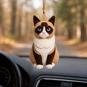 May include: A felt cat car charm with a grumpy expression. The charm features a brown and tan color scheme with blue eyes and a white chest. It hangs from a brown string, suspended in front of a car's dashboard.