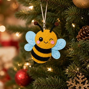 May include: A felt bee ornament featuring a yellow and black striped body, light blue wings with stitched details, and a winking face with pink cheeks. The bee has two black antennae with brown tips and hangs from a white string with a metal clasp.